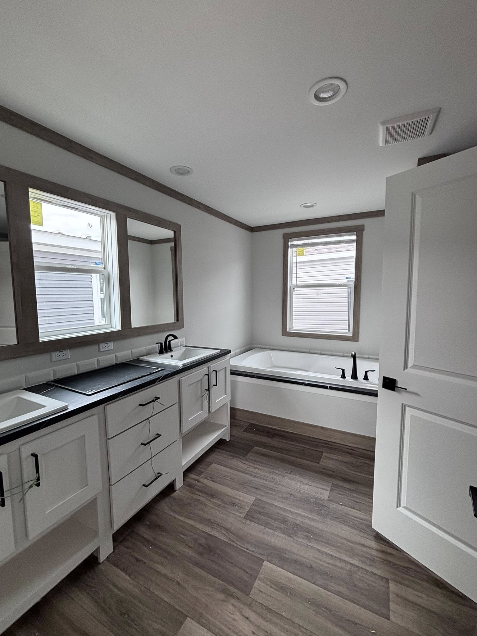 This extensive image gallery showcases various models from the "Boujee XL" series, including luxury tiny homes, park models, and spacious manufactured residences that blend a modern farmhouse aesthetic with high-end functional design. The exteriors are characterized by high-contrast color schemes, featuring white or cream board-and-batten siding paired with dark charcoal gables, gabled front porches with black or white support pillars, and durable metal roofing. Inside, the living areas emphasize an upscale, open-concept lifestyle with vaulted ceilings—some featuring natural wood planks or exposed beams—complemented by wide-plank wood-look flooring and crisp shiplap or neutral-toned walls. Gourmet kitchens are designed for high functionality, offering cabinetry in finishes ranging from light natural oak to elegant black, paired with marble-patterned or wood-block countertops, stainless steel farmhouse sinks, and large center islands with modern pendant lighting. Additional luxury elements include integrated booth seating, transom windows for maximum natural light, industrial-style ceiling fans, and spacious bedrooms with neutral carpeting. Contemporary bathrooms complete the homes with white vanities, black hardware, and choices of walk-in showers or large soaking tubs.