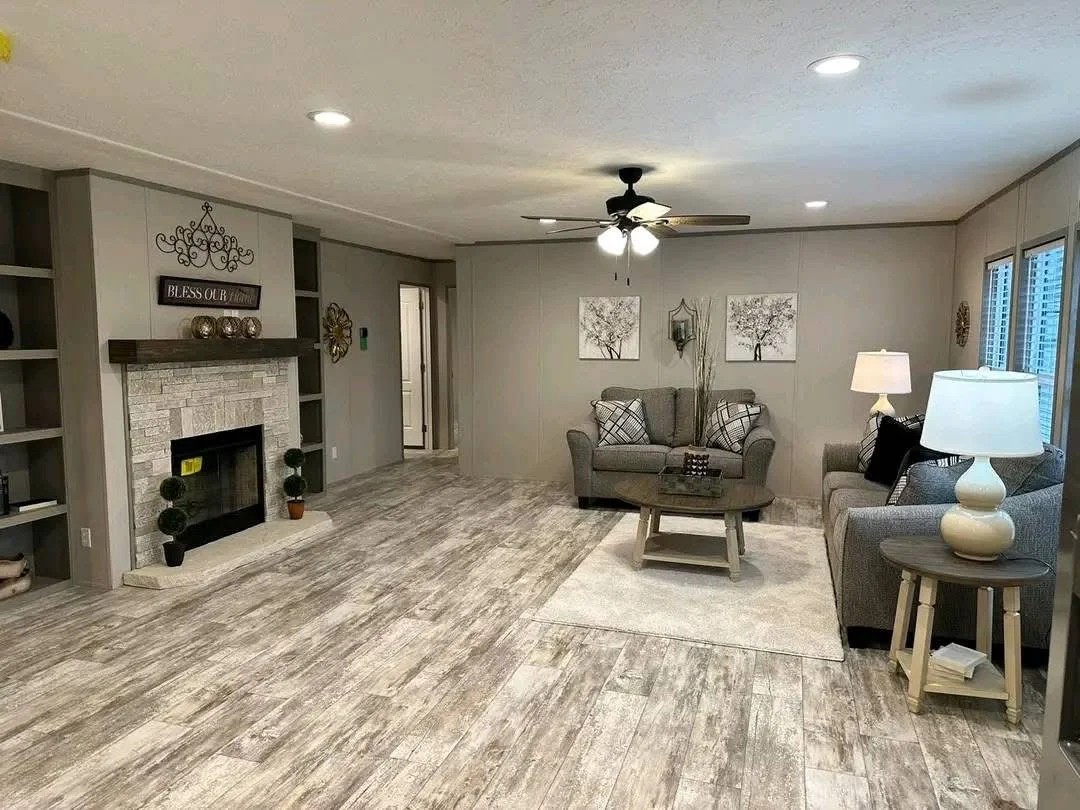 "A spacious, open-concept living room featuring grey wood-look flooring and neutral-toned walls. The room includes a grey fabric sofa and loveseat set, a round dark wood coffee table on a cream area rug, and a stone-look fireplace with a dark wood mantel and flanking built-in bookshelves. The space is illuminated by recessed lighting and a central ceiling fan."