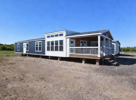 Modern two-story tiny home on wheels with blue and white exterior featuring expansive wraparound porch with white railings and wooden columns, gabled metal roof, large grid-pattern windows for maximum natural light, elevated deck on trailer chassis with concrete block foundation, spacious front entry staircase with handrails, dual-tone shiplap siding in navy blue and crisp white, bright sunny day setting ideal for mobile tiny house living, off-grid adventures, affordable modular housing, or vacation cabin retreat.