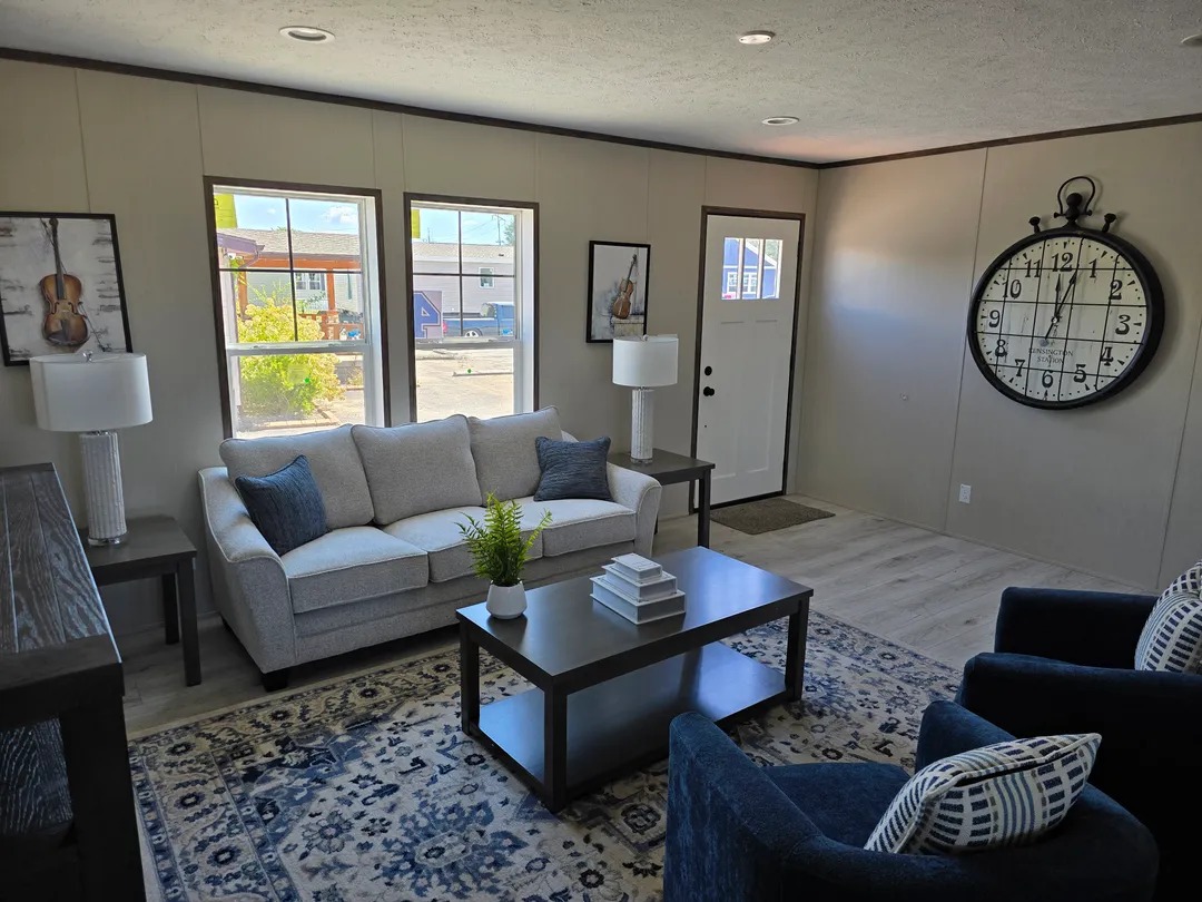 Cozy living room with a light gray sofa, blue armchairs, and a large wall clock. Sunlight streams through three windows, creating a warm ambiance.