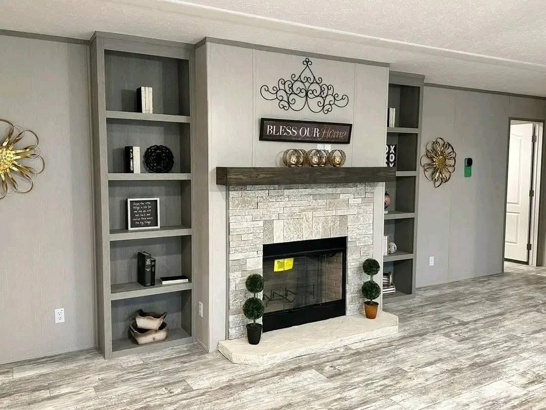 A modern living room feature wall with a stone-look fireplace and a dark wood mantel. The fireplace is flanked by tall, built-in grey bookshelves. Above the mantel is a 'Bless Our Home' sign and decorative metal scrollwork. The room has grey wood-look flooring and neutral-toned paneled walls."