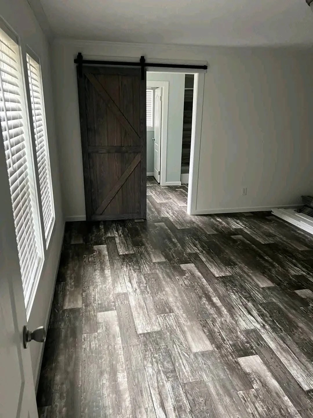 Room with rustic wood flooring, white walls, and a dark wooden sliding barn door partially open, revealing a bright adjacent room.