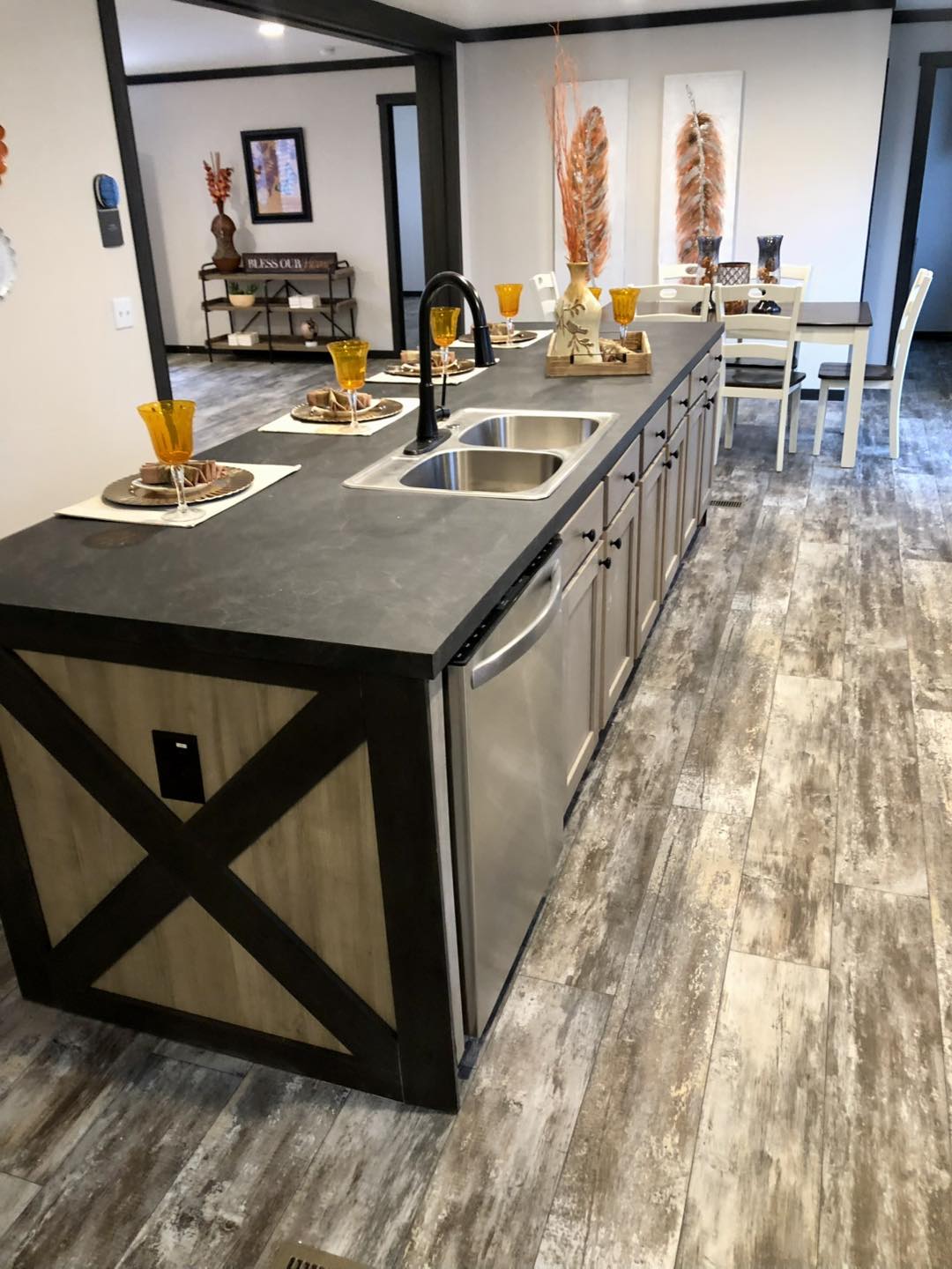 Luxury modern manufactured home showcase featuring premium double-wide floor plans with diverse exterior and interior designs. Exteriors include a white board-and-batten APH 1662C model and a sleek dark charcoal mobile home with rustic wood shutters and a white brick-patterned foundation. Interior highlights feature open-concept living rooms with integrated electric fireplaces, shiplap accent walls, and vaulted ceilings with rustic wood beams. Gourmet kitchens are equipped with oversized marble-look or charcoal waterfall islands, farmhouse cabinetry, and custom pull-out pantry storage. The master suites boast bold black accent walls and cowhide rugs, while spa-inspired bathrooms include freestanding soaking tubs, walk-in tile showers with black trim, and LED-mirrored double vanities. Additional features include a modern laundry room with marble-look countertops and a spacious covered front porch with a dark wood-plank ceiling and a signature hanging wicker egg chair.