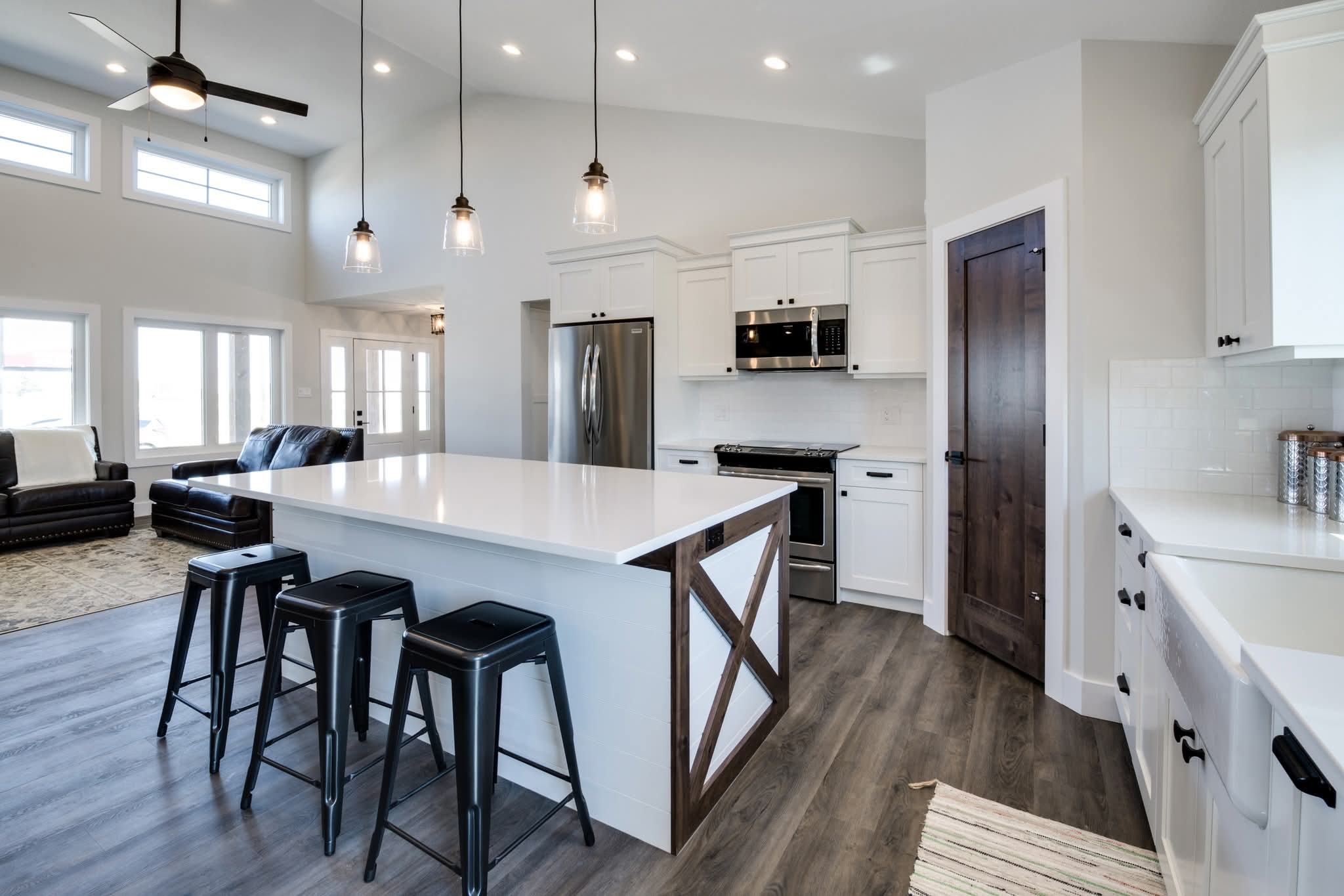 Spacious modern kitchen with a large white island, dark stools, and stainless steel appliances. High ceiling with pendant lights, wood accents, and plank flooring create a cozy, inviting atmosphere.