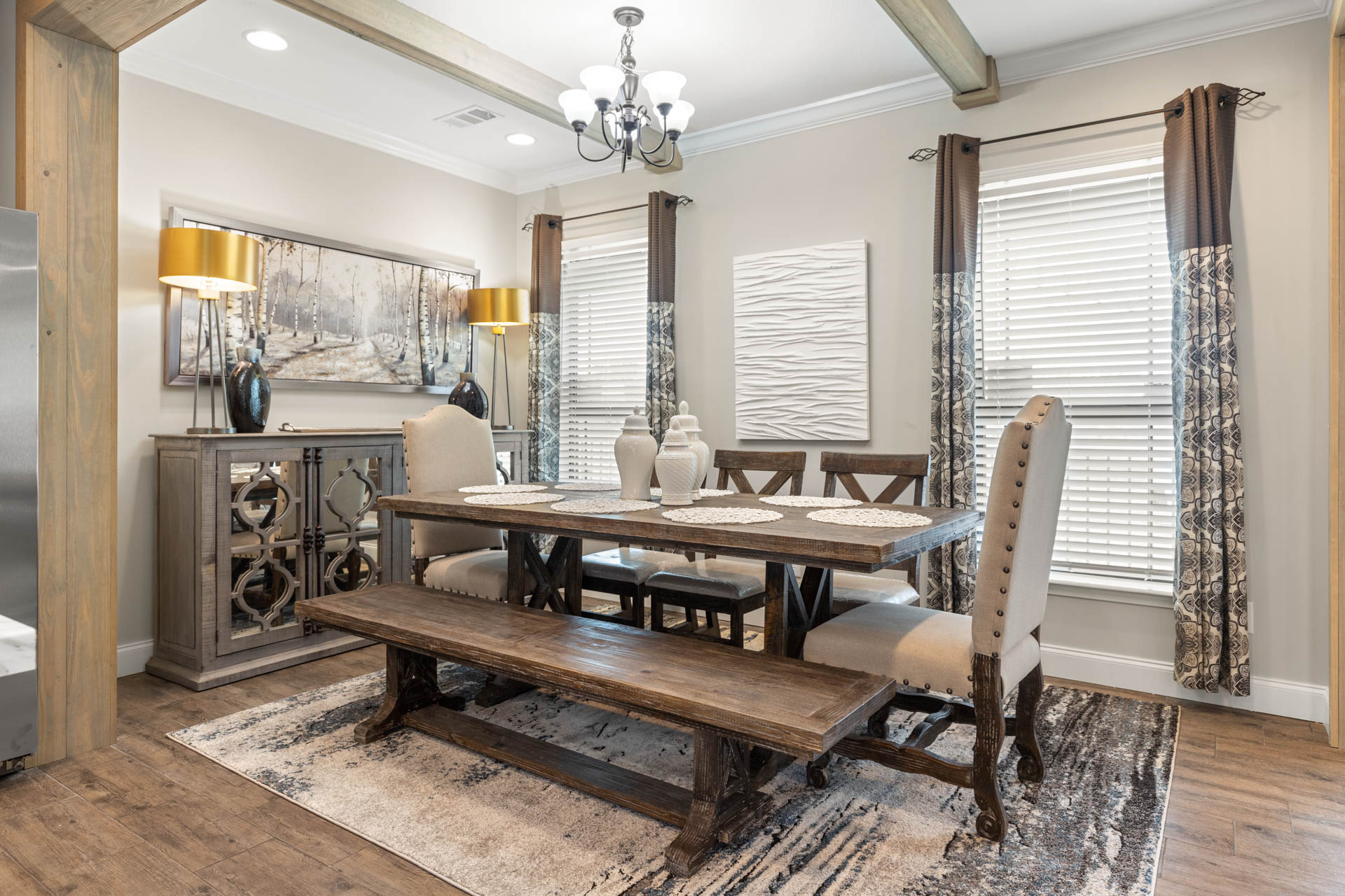 Warm dining room with rustic wood table and bench, upholstered chairs, and chic decor. Large windows with patterned curtains brighten the space.