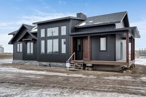 Modern black modular home on a snowy field, featuring large windows and a wooden porch. The setting feels cold yet contemporary and inviting.