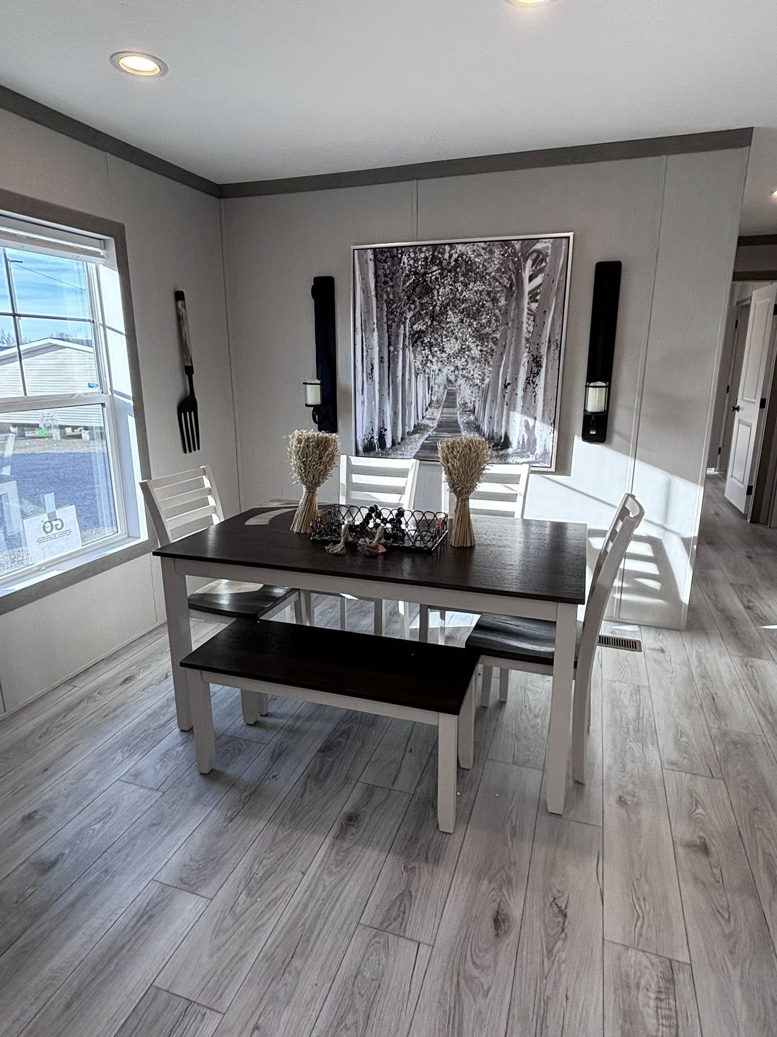 A modern farmhouse-style dining area featuring a dark wood-top table with white legs, a matching dark wood bench, and three white wooden chairs. On the table sits a decorative wire basket and two wheat stalk arrangements. The wall behind is decorated with a large black-and-white canvas of a forest path, flanked by two black wall sconces and a decorative wooden fork. The room has light grey wood-plank flooring and a large window to the left.