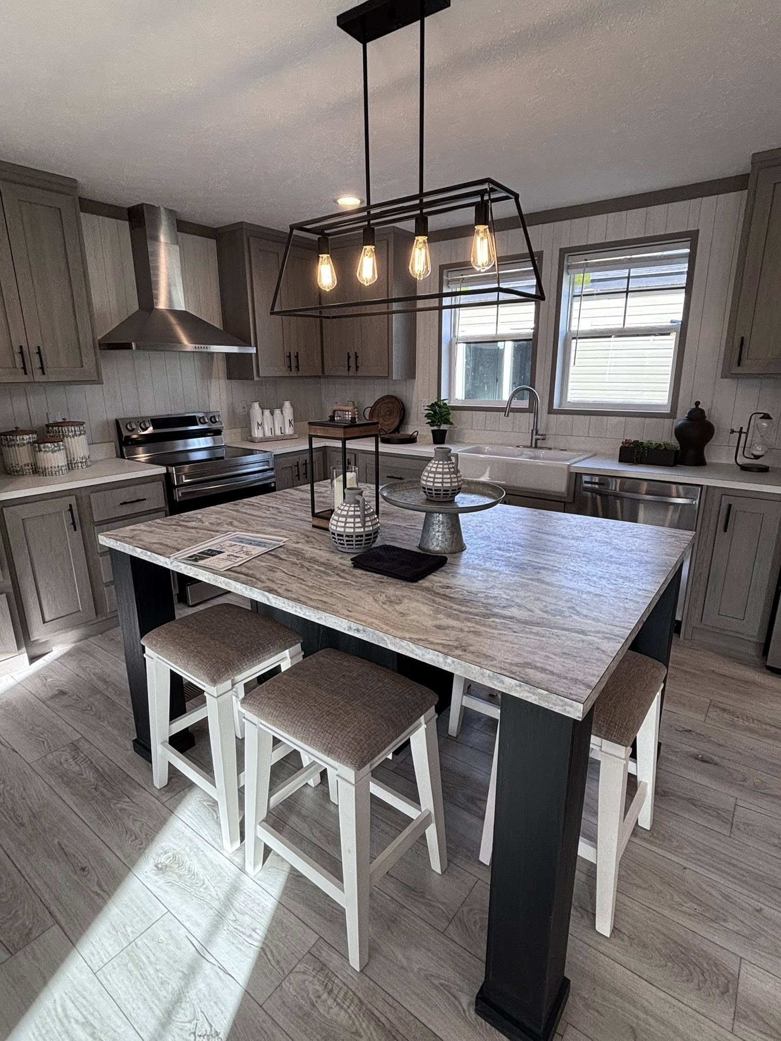 A rustic-modern dining set featuring a dark wood-top table with white legs, paired with three matching white wooden chairs and a long dark wood bench. The table is decorated with two wheat stalk bundles and a metal wire basket. On the wall hangs a large black-and-white canvas of a tree-lined path, flanked by two black candle sconces, while a large decorative wooden fork hangs to the left.
