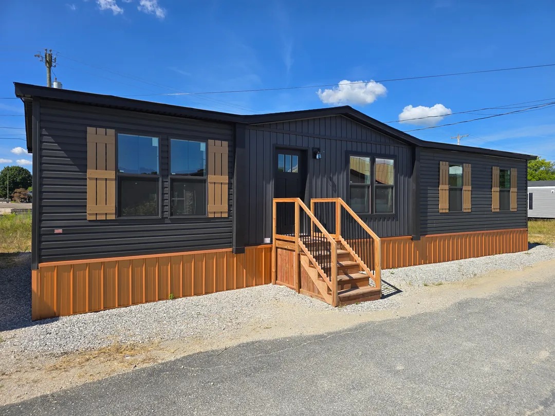 Modern black and brown modular home with wooden shutters and small front steps. Set on gravel under a bright blue sky with sparse clouds.