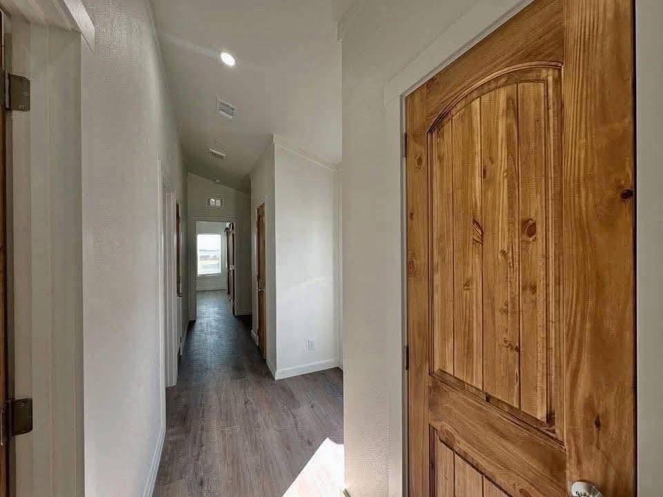 Narrow, well-lit hallway with wooden doors and light-colored walls. Wooden flooring leads to a window with a view, conveying a warm, inviting ambiance.