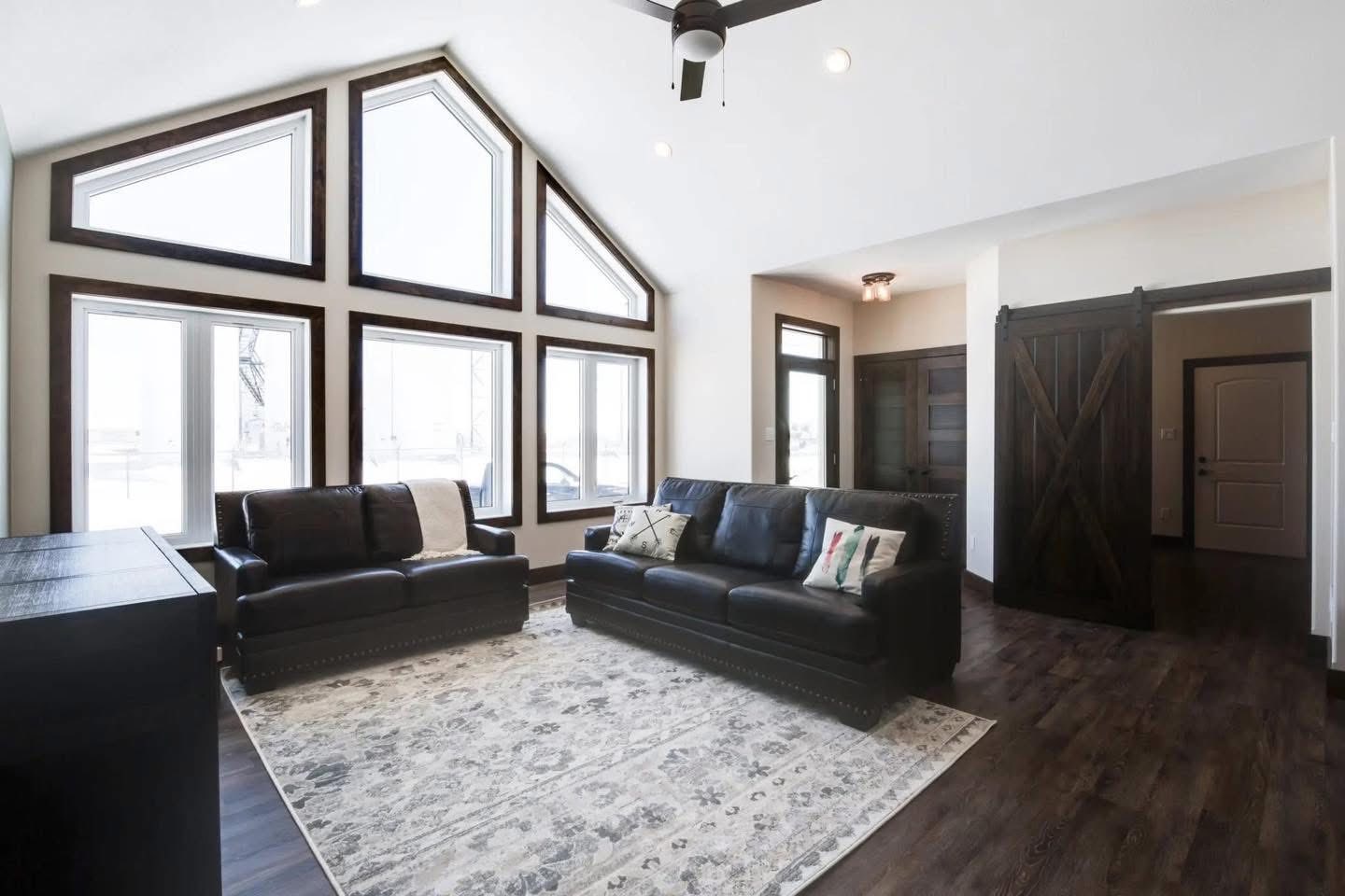 A bright living room features a high ceiling, large triangular windows, two black leather sofas, a patterned rug, dark wooden flooring, and a barn door.