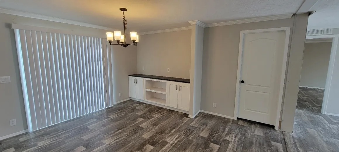 A spacious room with wooden flooring, a modern chandelier, and vertical blinds covering a large window. White cabinets and a closed door are present.