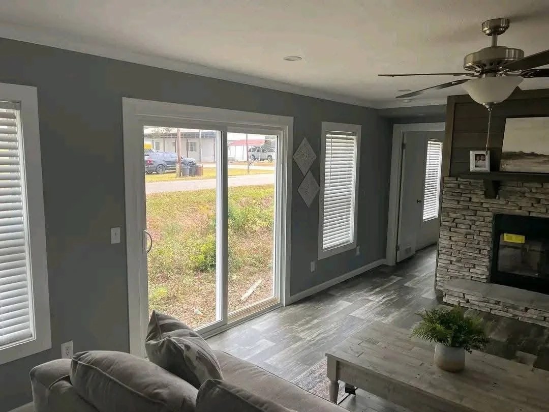 A cozy living room with gray walls, a stone fireplace, and large windows. Light floods the room, highlighting a gray couch and wooden coffee table.