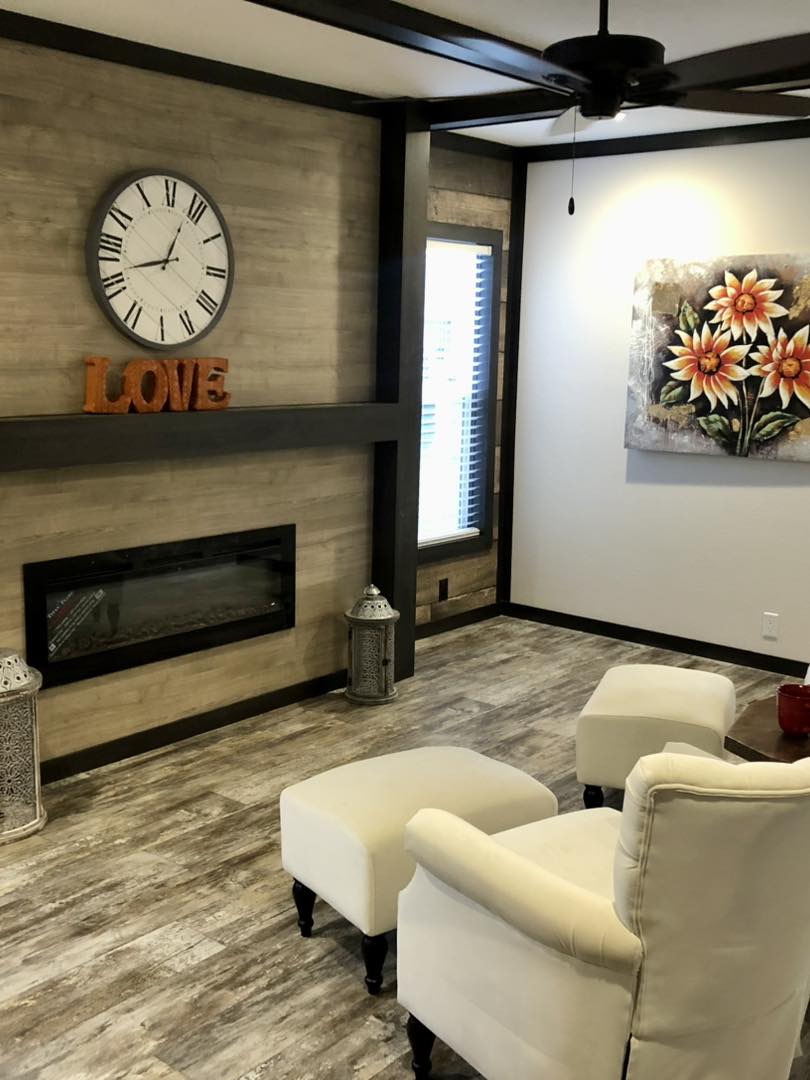 Luxury modular and manufactured home showcase featuring premium floor plans like the APH 1662C. Exteriors display white board-and-batten farmhouse styles and dark charcoal siding with rustic wood shutters. High-end interior highlights include open-concept living rooms with integrated electric fireplaces, shiplap accent walls, and vaulted ceilings with dark wood beams. Gourmet kitchens feature large marble-look waterfall islands, herringbone tile backsplashes, and farmhouse-style cabinetry with custom pull-out pantry storage. Additional luxury features include a master suite with a bold black accent wall, a spa-like bathroom with a freestanding soaking tub and LED-mirrored double vanity, a modern laundry room with marble-look countertops, and a tranquil covered front porch with a wood-plank ceiling and a stylish hanging wicker egg chair.