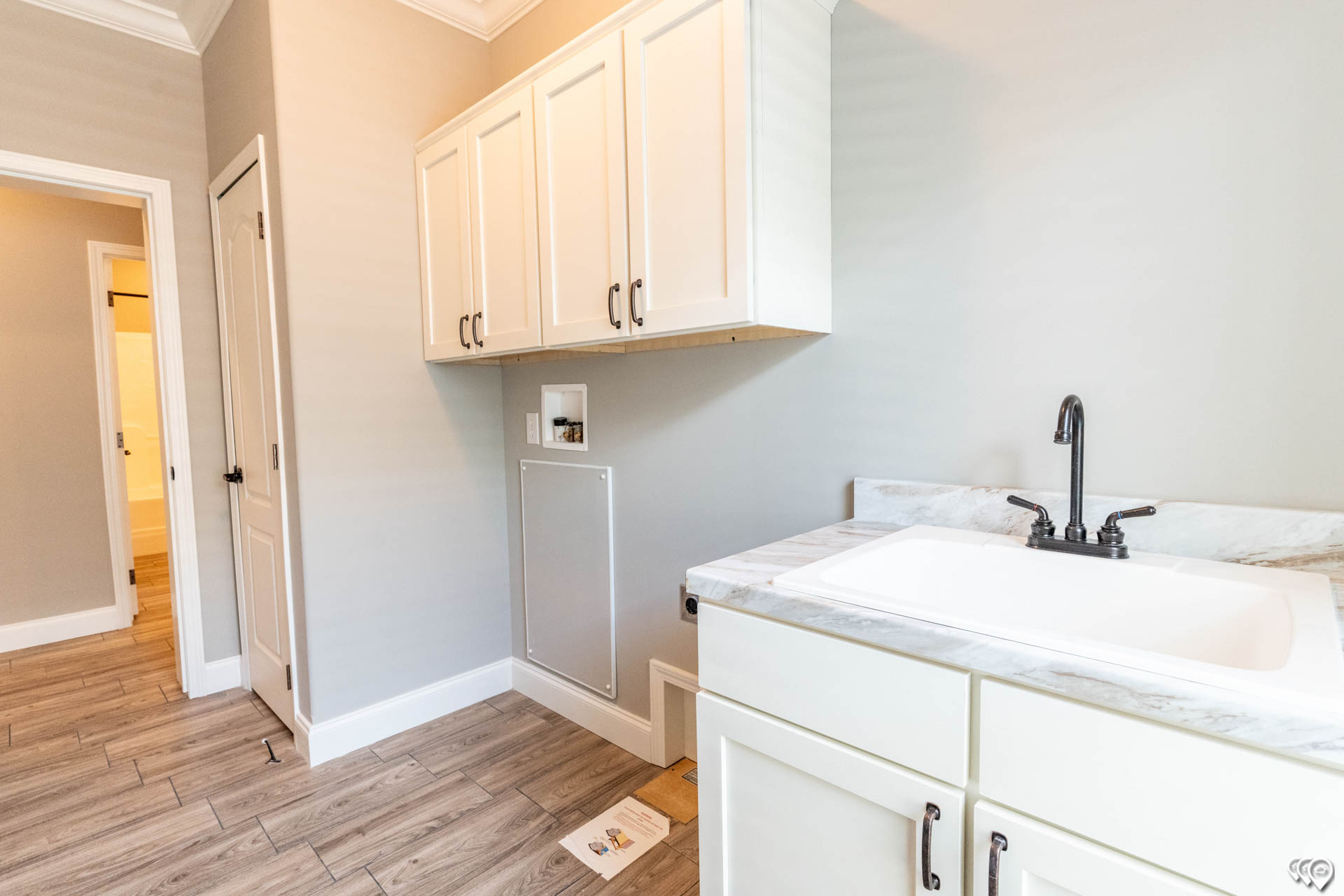 A laundry room with gray walls and wood-like tile flooring. It features white cabinets, a marble countertop with a black faucet, and an open doorway.
