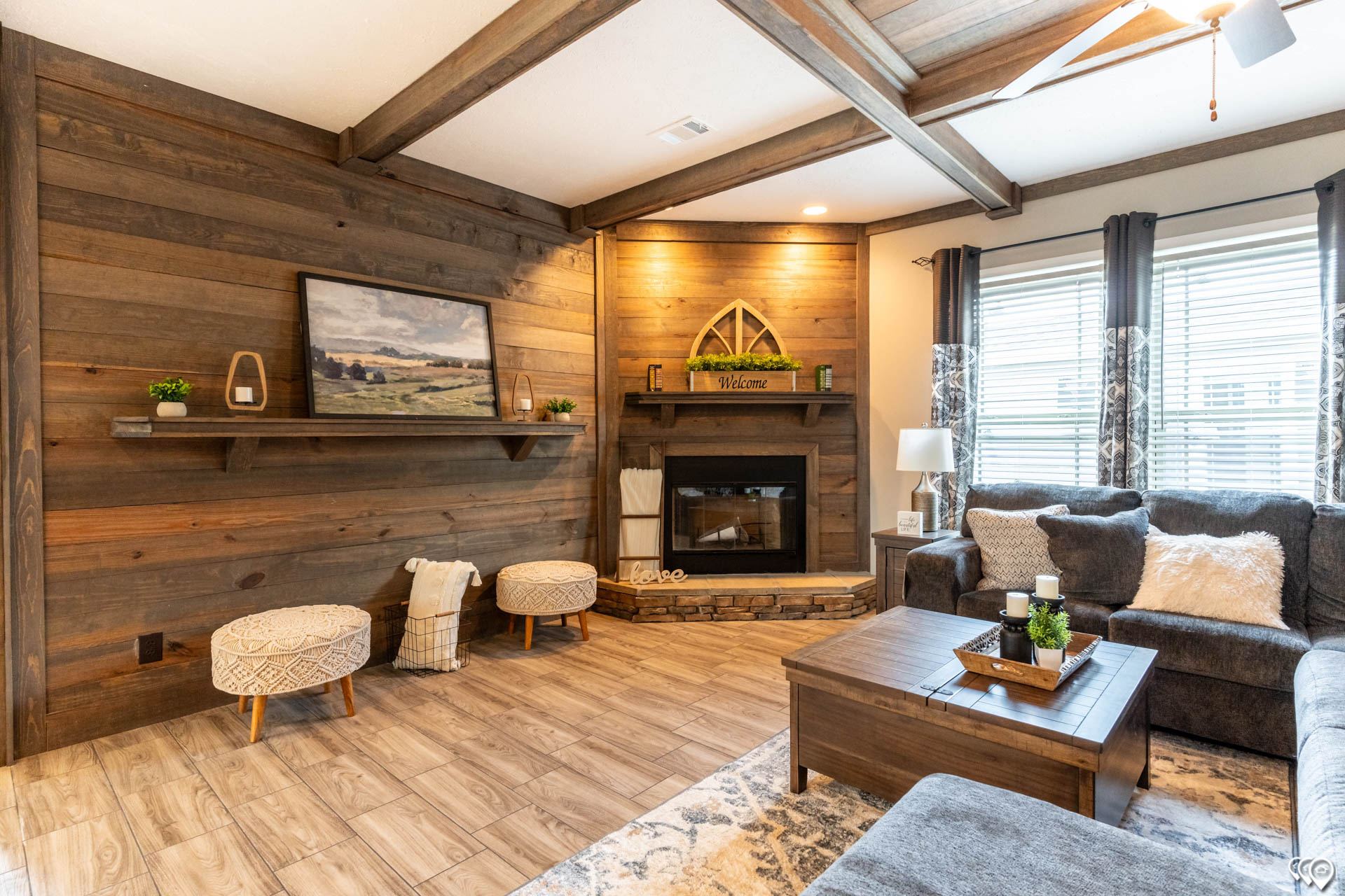 Cozy living room with wood-paneled walls and ceiling beams. Features a fireplace, gray sofas, cushioned stools, and a wooden coffee table. Natural light from large windows adds warmth.