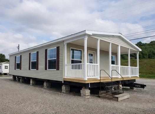 Manufactured home on blocks in a gravel lot, featuring a small front porch with white railing and steps, set against a cloudy sky and trees.