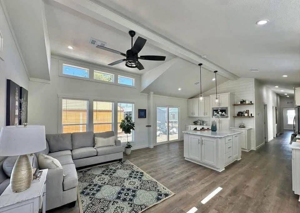 Modern open-concept living area with white walls, high ceilings, and a ceiling fan. Features a cozy gray sofa, patterned rug, and a bright kitchen with an island. Natural light streams through large windows, creating an inviting atmosphere.