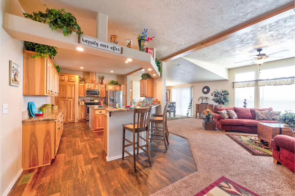 Open-concept ranch-style home interior featuring a rustic kitchen with light wood cabinetry, granite countertops, a breakfast bar with wooden stools, and an integrated living room area with hardwood and carpet flooring.