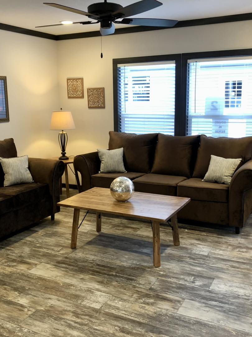 High-end manufactured home showcase featuring premium double-wide floor plans, including the white board-and-batten APH 1662C and dark charcoal models with rustic wood shutters. Luxury interior highlights feature open-concept living rooms with integrated electric fireplaces and shiplap accent walls, gourmet kitchens with oversized marble-look or charcoal islands and farmhouse-style cabinetry, and custom laundry rooms with ample storage. The modern design is elevated by vaulted ceilings with dark wood beams, master suites with bold black accent walls and cowhide rugs, luxury bathrooms featuring freestanding soaking tubs and LED-mirrored double vanities, and cozy guest bedrooms with white iron bed frames. Outdoor living is showcased by a spacious covered front porch with a dark wood-plank ceiling and a signature hanging wicker egg chair.