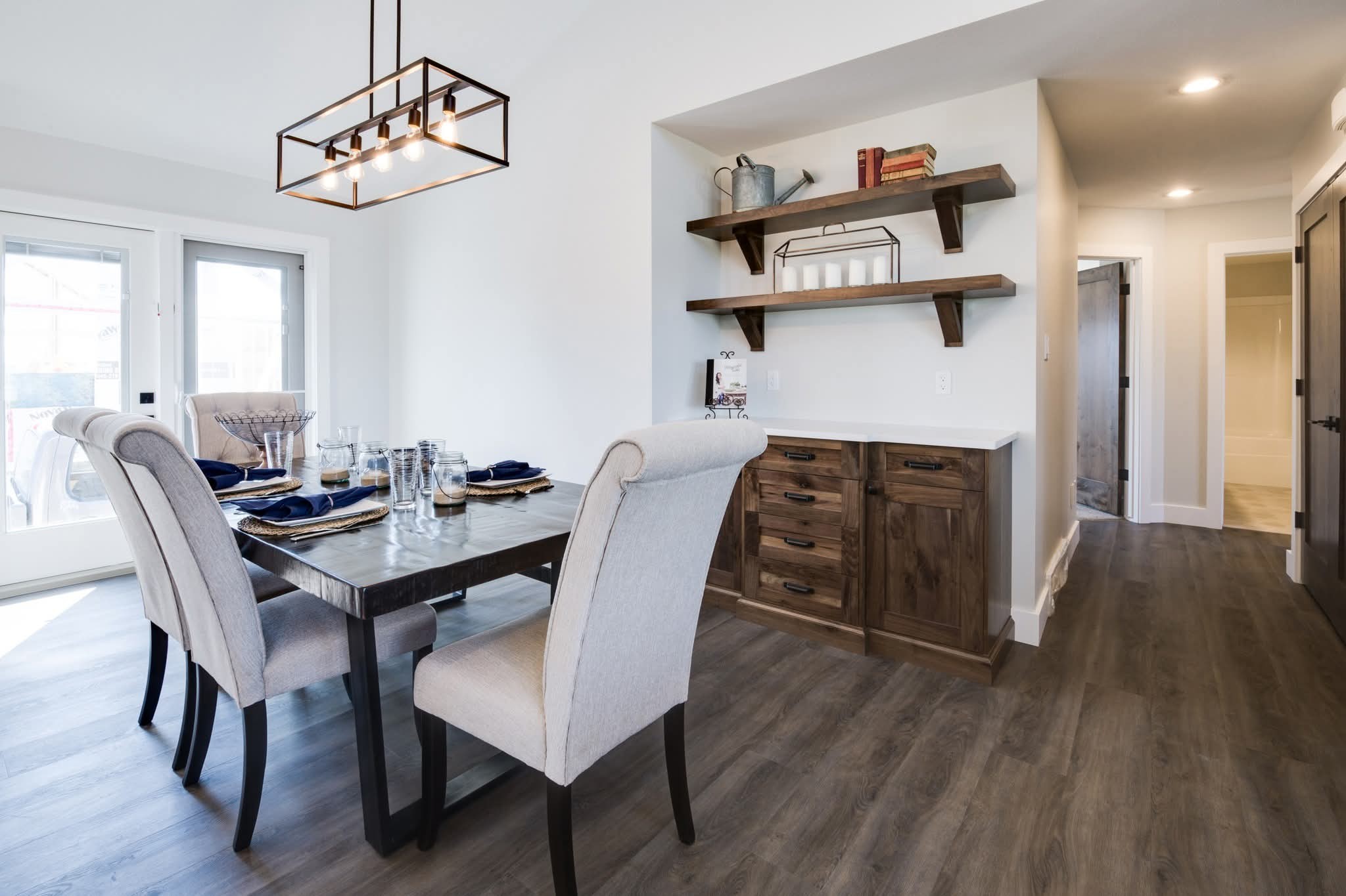 A modern dining room with a wooden table set for four, cushioned chairs, and a sleek rectangular chandelier. Wooden shelves and cabinets add warmth.
