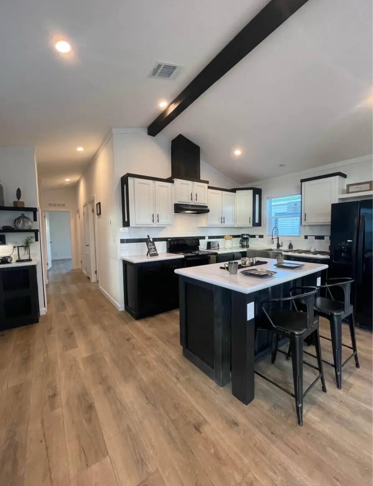 Modern kitchen with black and white cabinetry, a central island with stools, and wooden flooring. Bright, clean space with recessed lighting.