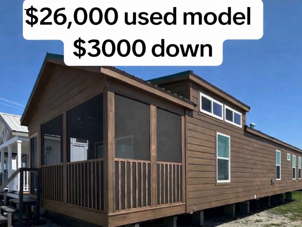 Brown manufactured home with a screened porch, set on a clear day. Text reads "$26,000 used model" and "$3,000 down." Affordable housing option.