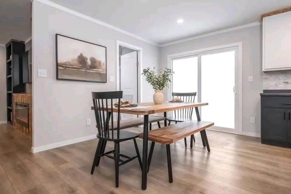 A modern dining area with a wooden table, two black chairs, and a bench. A vase with greenery is on the table. Bright and minimalist decor.