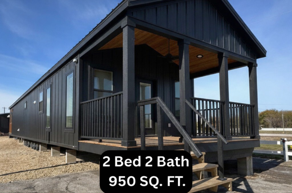 Modern black tiny home with a small porch, situated in a rural area under a clear blue sky. Text overlay: "2 Bed 2 Bath, 950 SQ. FT."