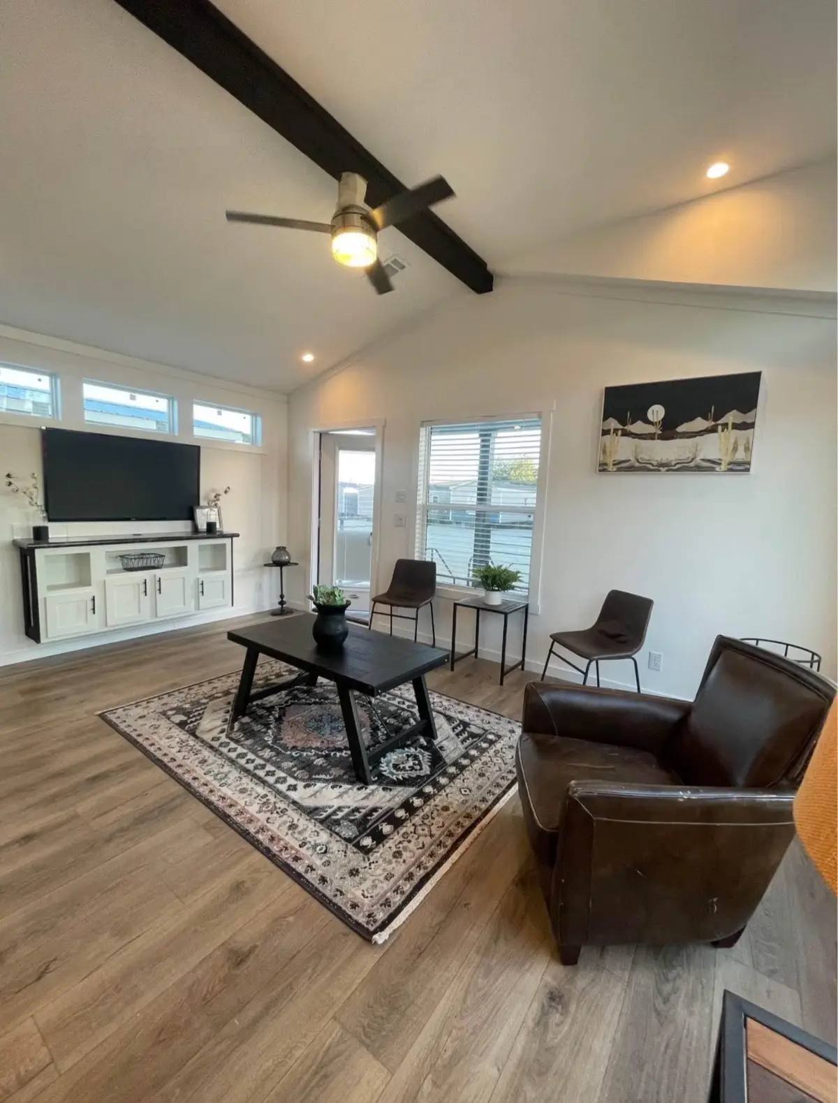 Spacious living room with wooden floors, featuring a ceiling fan, black coffee table on an ornate rug, brown chairs, and modern decor under soft lighting.