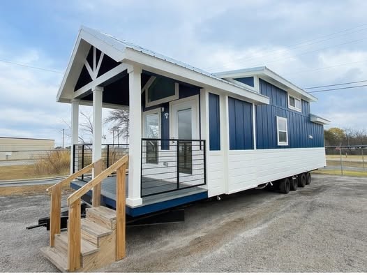 A modern tiny house on wheels with a blue and white exterior is parked on a gravel lot. It features a wooden porch with steps, under a cloudy sky.