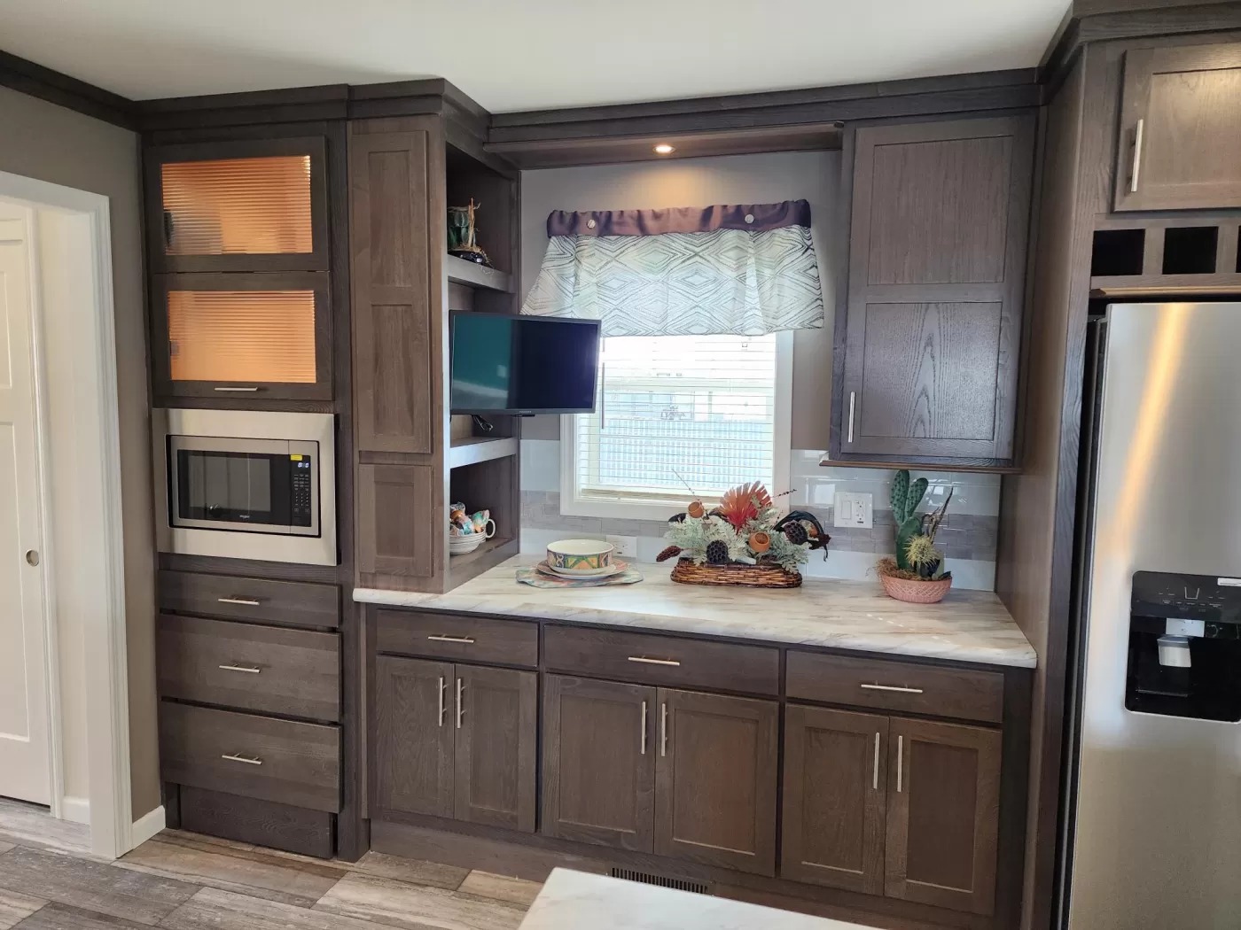 A modern kitchen with dark wood cabinets, a built-in microwave, and a small TV. Countertop displays a flower arrangement and potted plant, under a window with a patterned curtain.