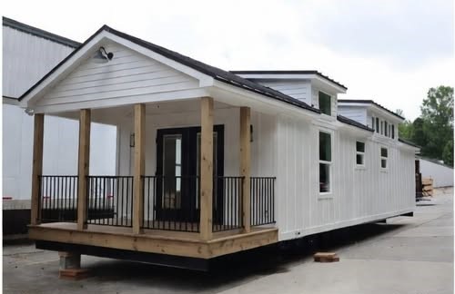A white tiny house with a gabled roof and front porch sits on a concrete lot. It features a black door, large windows, and wooden columns, conveying simplicity.