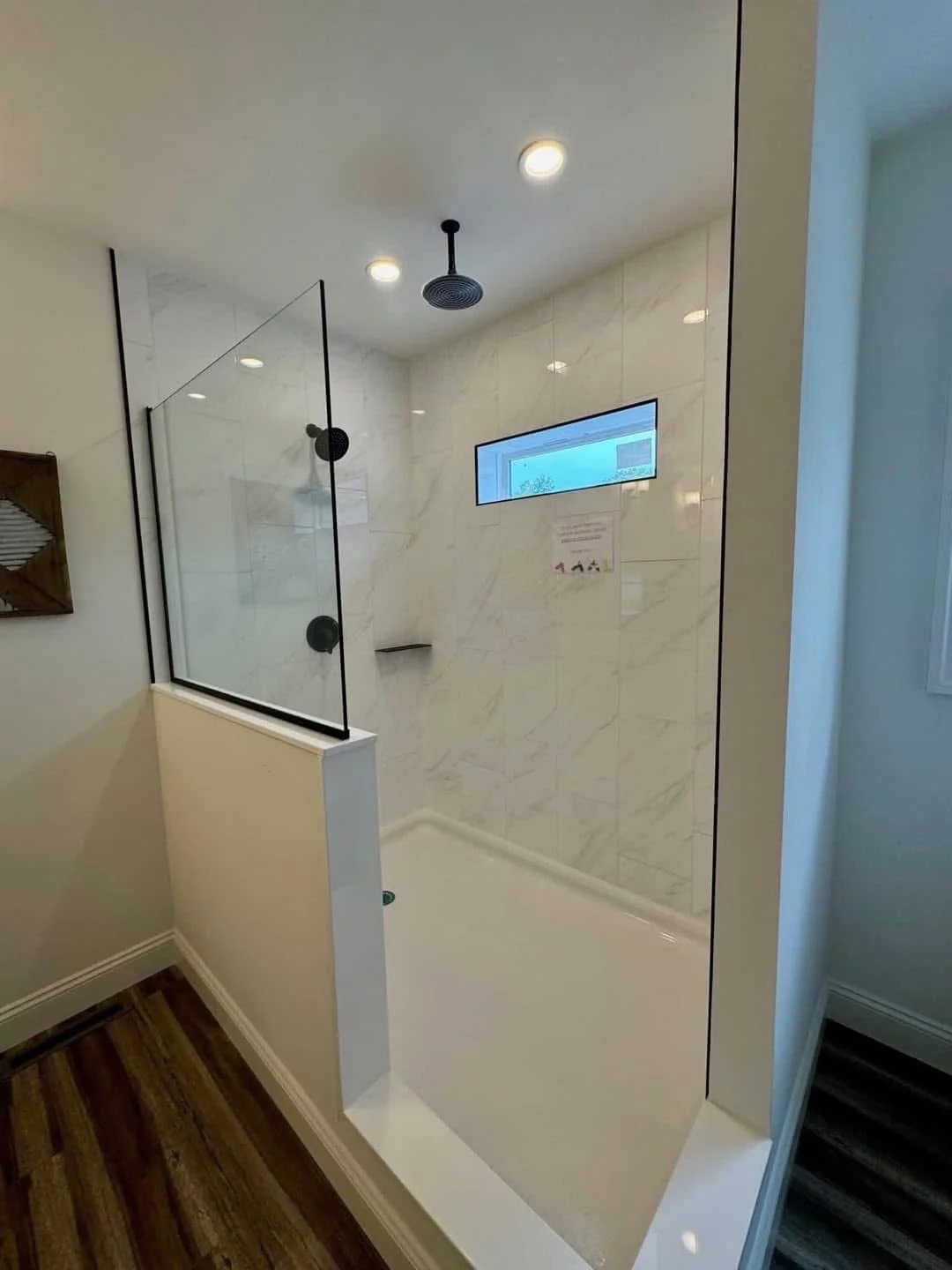 Modern bathroom shower with glass enclosure, black fixtures, and white tile walls. A rectangular window adds natural light. Hardwood flooring outside the shower.
