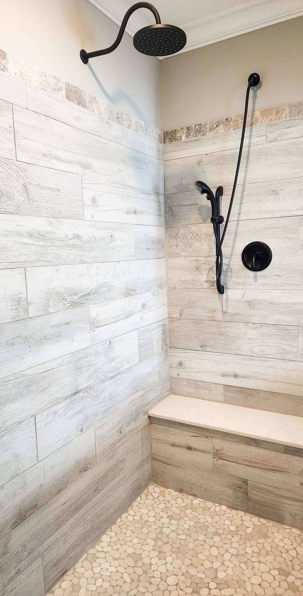 Modern shower with light wood-style tiles and pebble stone floor. Features a black rain showerhead, handheld shower, and built-in bench.