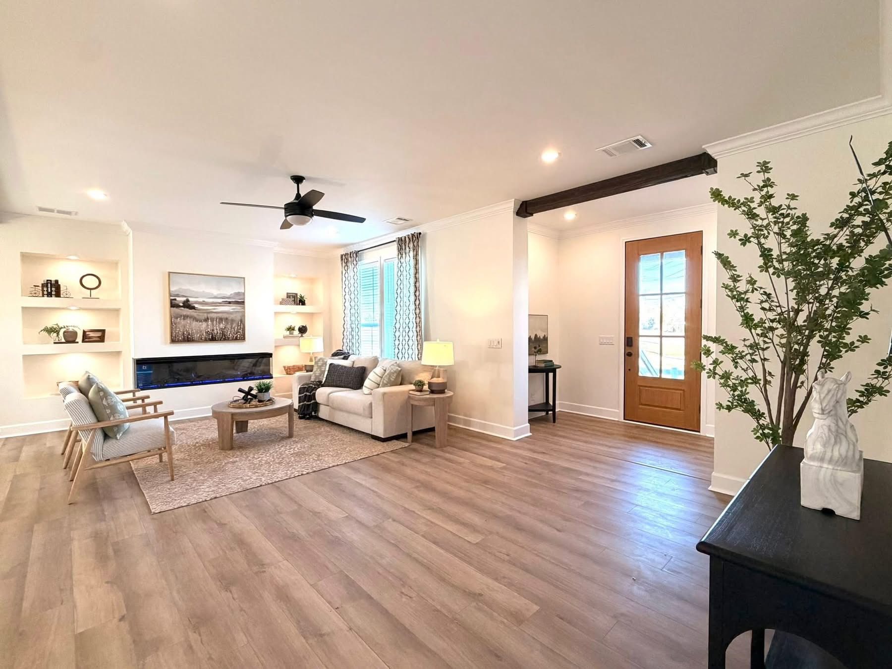 Spacious living room with light wood floors, white walls, and a ceiling fan. Features a white couch, wooden furniture, and large windows. Cozy and inviting atmosphere.