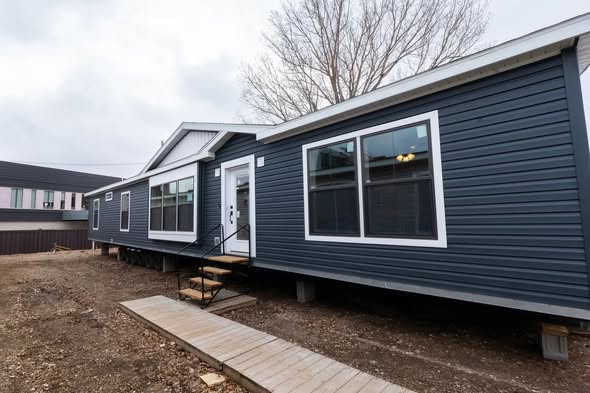 A modern, dark blue mobile home with large windows sits on a dirt plot. A wooden ramp leads to the entrance. The scene is overcast, with leafless trees.