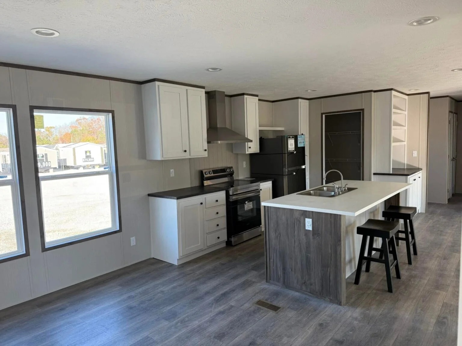 Modern kitchen with light wood floors, white cabinets, black appliances, and a central island with stools. Large windows provide natural light.