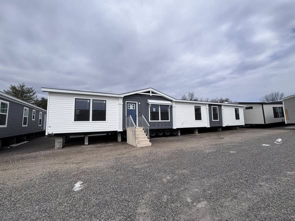 This extensive image gallery showcases various luxury models from the "Boujee XL" series, including tiny homes, park models, and manufactured homes that blend modern farmhouse style with high-end functional design. The exteriors are characterized by crisp white, cream, or grey board-and-batten siding accented by charcoal gables, spacious gabled front porches with black or white pillars, and durable metal roofs in white or dark grey. Interior living spaces feature a sophisticated open-concept layout with vaulted ceilings, exposed natural or painted wood beams, white shiplap walls, and wide-plank wood-look flooring. Gourmet kitchen and dining areas range from warm natural wood cabinetry with built-in booth seating to contemporary kitchens with white or black cabinets, marble-patterned countertops, stainless steel farmhouse sinks, and even vibrant teal appliances. Premium details are integrated throughout, including lofted sleeping areas with black metal railings and wooden ladders, industrial-style ceiling fans, transom windows for natural lighting, and modern bathrooms featuring walk-in marble-patterned tile showers and generous vanity storage.