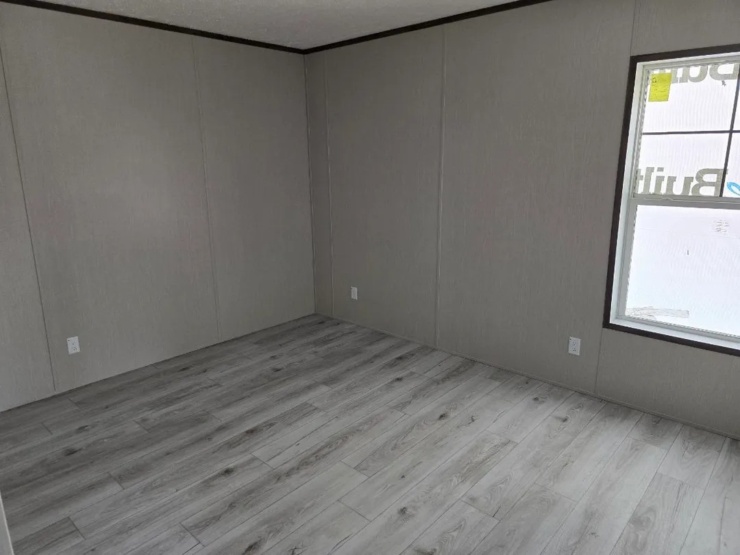 Empty room with light gray walls and polished wooden floor, featuring one window on the right side. Bright, minimalistic, and spacious atmosphere.