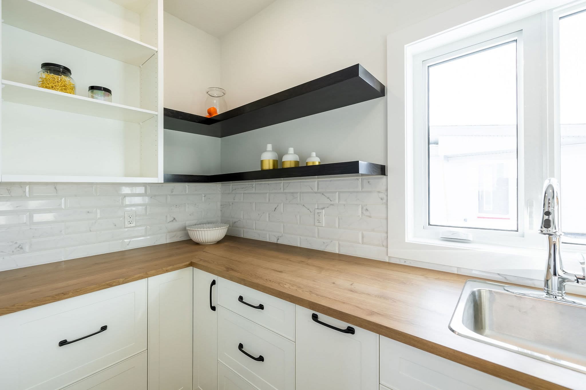 A modern kitchen corner with light wooden countertops and white cabinets. Open shelves hold jars and bottles. A window and stainless steel sink add brightness.