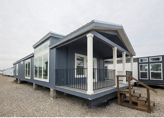 A blue manufactured home with large windows and a small front porch featuring steps and white columns, set on a gravel surface under a cloudy sky.