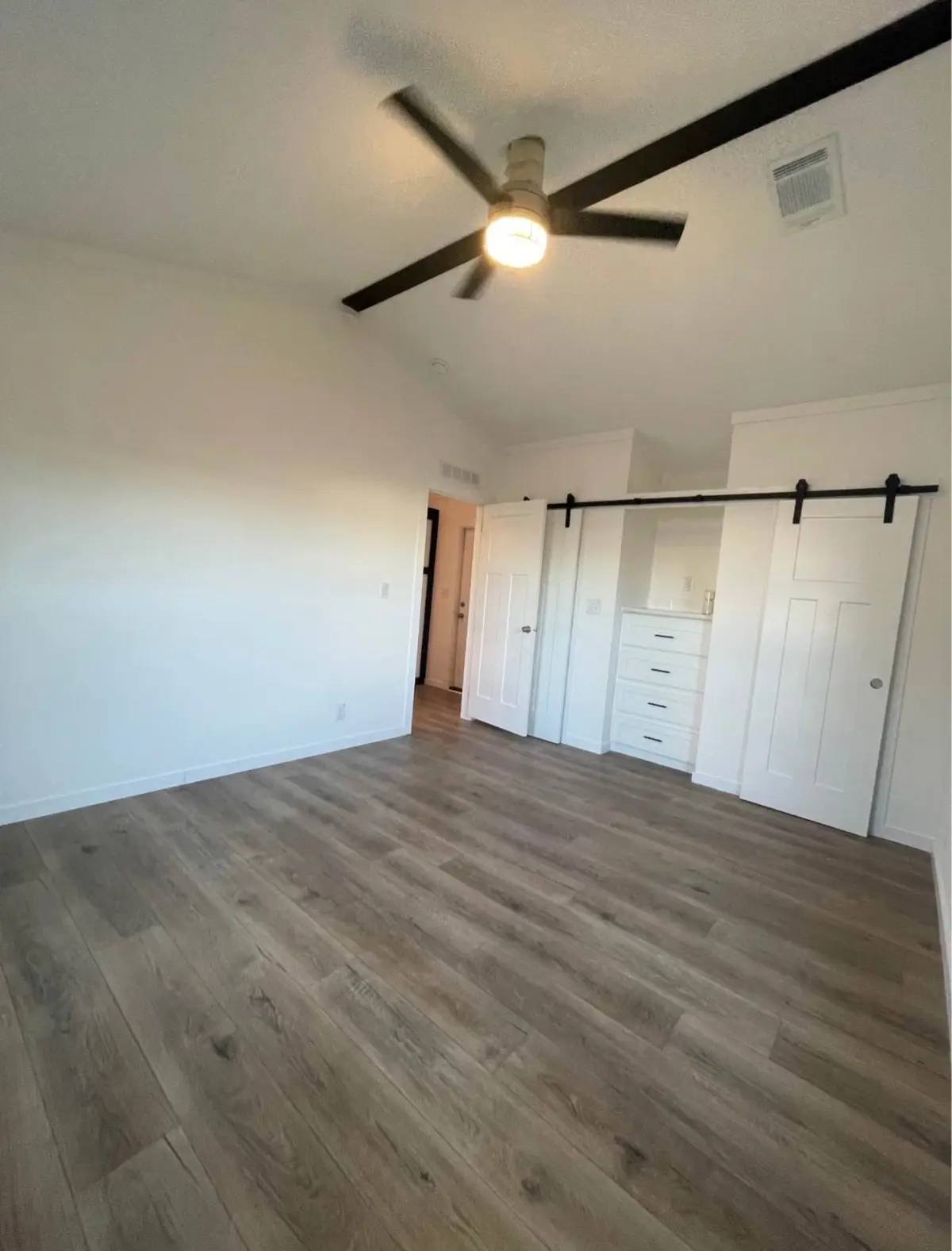 Spacious room with light wood flooring, a ceiling fan, and white walls. Features a sliding barn door closet with built-in drawers. Bright and modern.