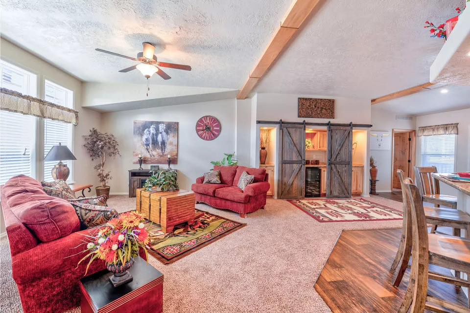 Open-concept ranch home interior featuring a rustic kitchen with light oak cabinets, granite countertops, and a breakfast bar, flowing into a dining room with a horse-themed rug and a spacious living room with vaulted ceilings, red sofas, and sliding barn doors.