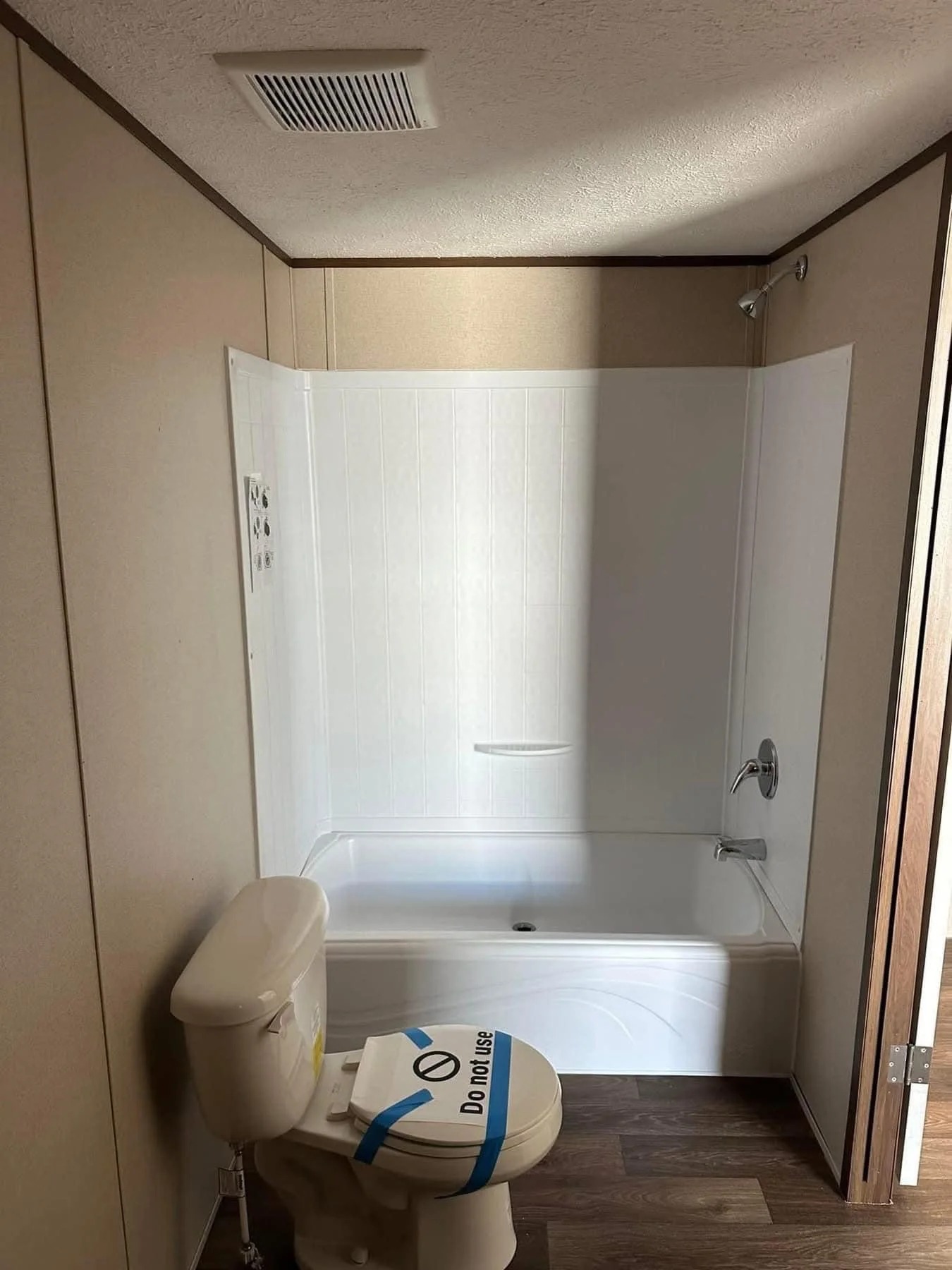 A small bathroom with beige walls, featuring a white bathtub and shower combo. In front is a toilet labeled with a "Do Not Use" sign in blue tape.
