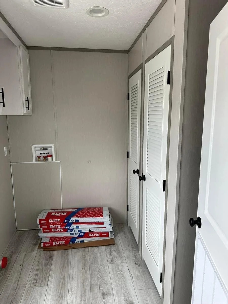 A small, unfurnished room with light gray walls and wood flooring features a stack of insulation packs labeled "Elite" beside white louvered closet doors.
