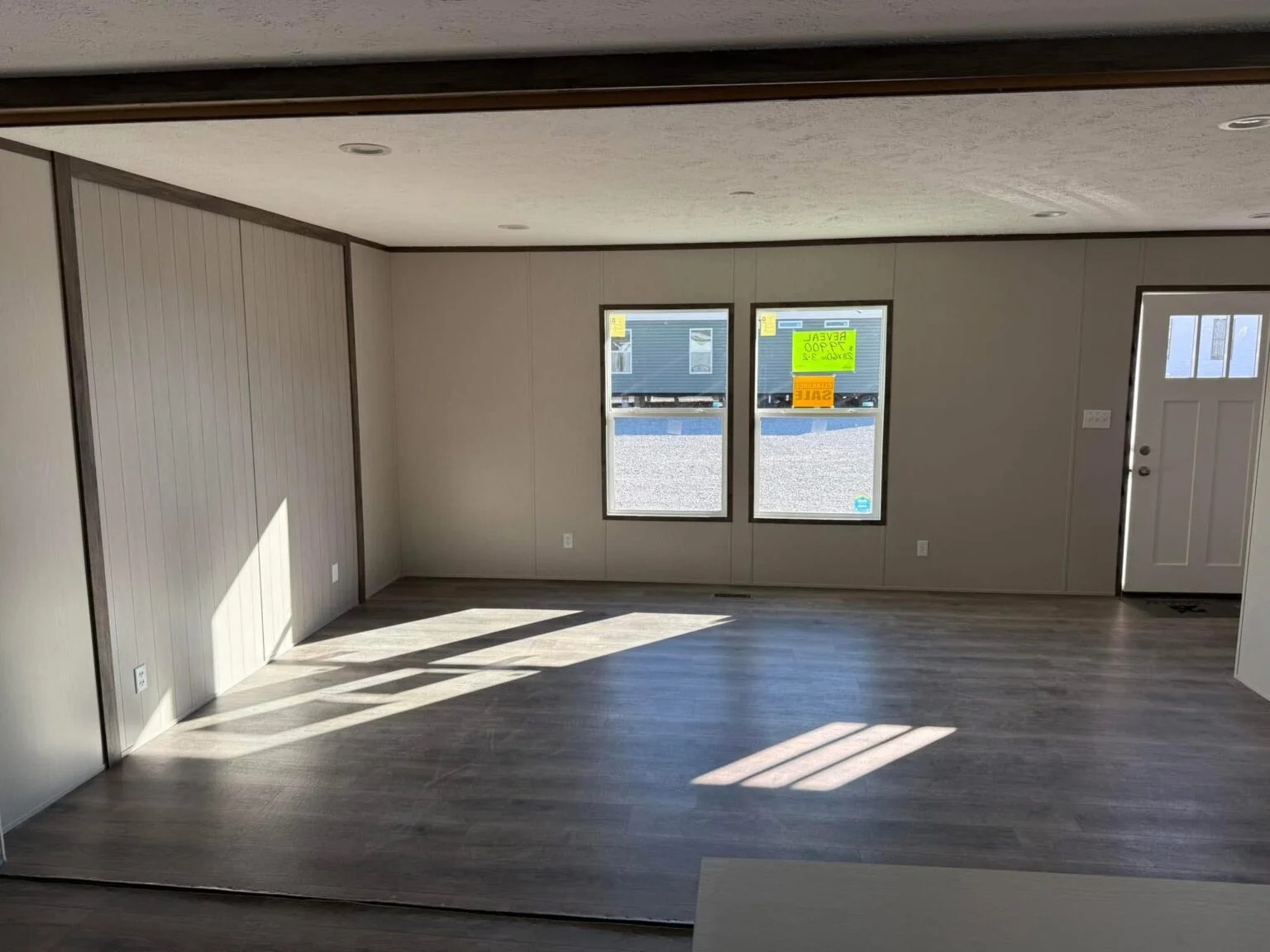 Spacious empty room with light gray walls and dark wood flooring. Two large windows and a door allow natural light to brighten the interior.