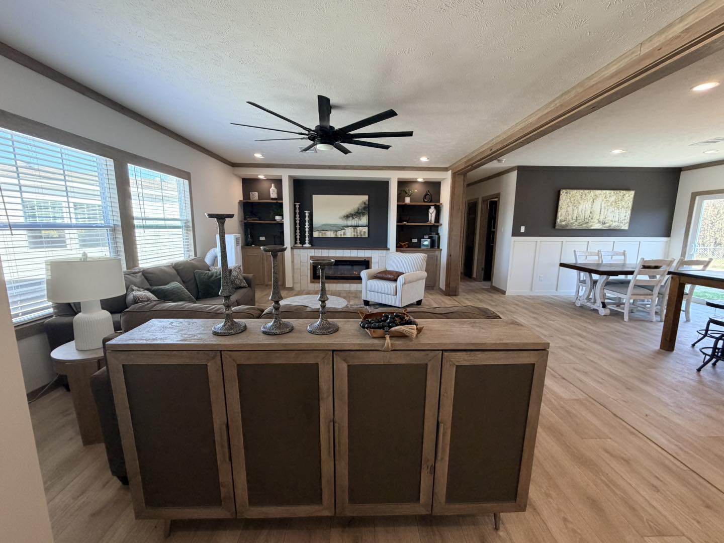 Modern open living space with light wood flooring. Features include a large ceiling fan, cozy seating area, dining table, and tall windows. Neutral tones create a calm atmosphere.