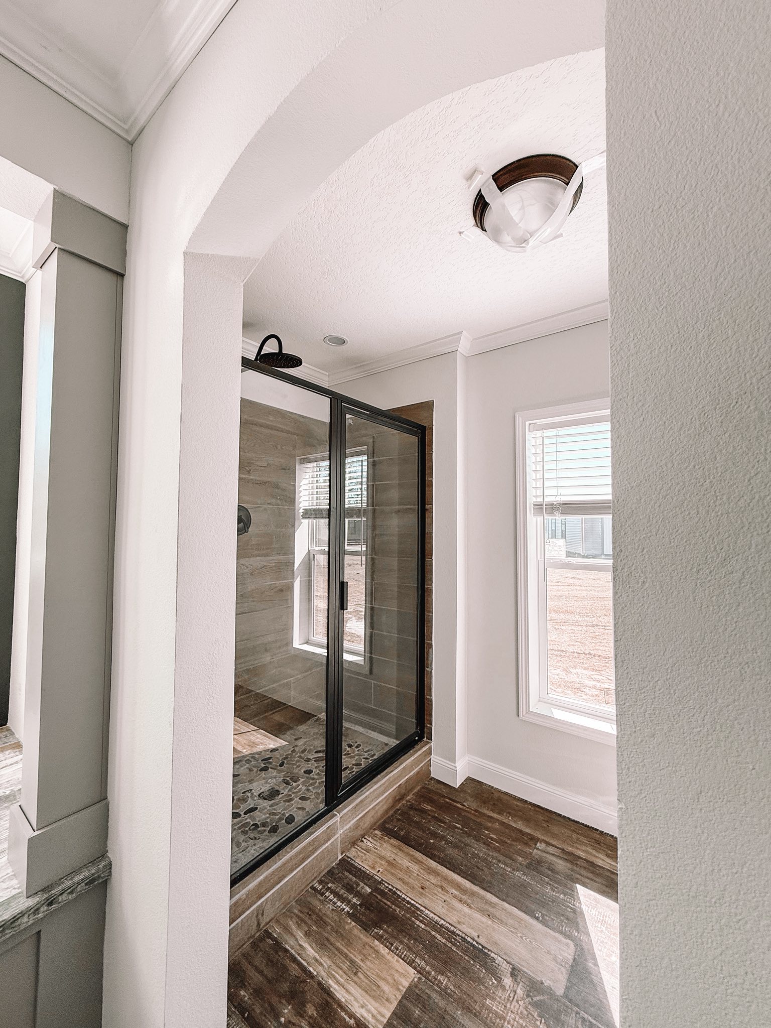 Bright bathroom with a wooden floor and a glass shower enclosure, illuminated by natural light from a large window. Modern and serene ambiance.