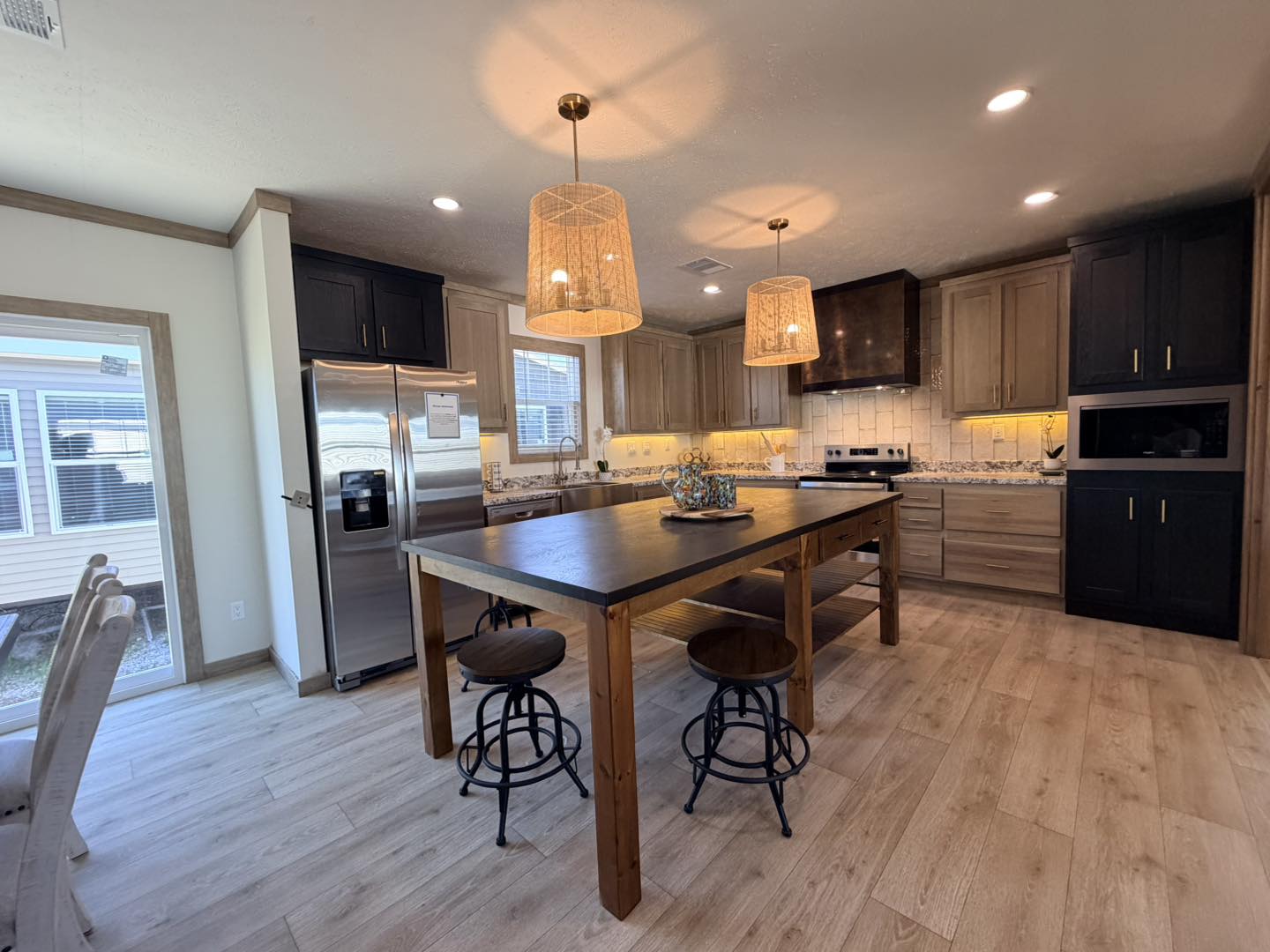 Spacious kitchen with a large wooden island, three stools, modern appliances, and warm pendant lights. Light wood floor and dark cabinets. Cozy ambiance.